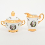 Porcelana Real, Mauá - SP(Fundada em 1943 por Fritz Erwin Schmidt).Um(01) açucareiro e uma(01) leiteira em porcelana nacional da década de 1950/60; esmaltação vítrea de tonalidade branca, corpo bojudo sobre base circular recortada, bico vertedor ondulado e revirado para fora, tampa abaulada encimada por puxador com feitio de fruto, asas laterais em grande voluta; decoração composta por reserva oval com cena galante de tradição rococó, representando casal em paisagem, associadas a ramagens florais esparsas aplicadas em estampagem policromada; bordas, bico vertedor, asas, friso superior e base integralmente recobertos por generosa douração. Marca da manufatura no verso. Medidas: Açucareiro: Alt.: 11,5 x Comp.: 13,5 x Larg.: 9,5 cm; Leiteira: Alt.: 10,5 x Comp.: 12 x Larg.: 7,5 cm.