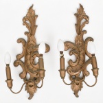 Par de arandelas de parede de dois lumes, em madeira entalhada e dourada, com braços aplicados em metal,  de estilo neorrococó, século XX; espaldar recortado e vazado com composição em volutas, folhas de acanto e elemento floral em relevo, remate superior com motivo vegetal, dois braços curvos por peça de secção tubular terminando em suportes cilíndricos para lâmpadas tipo vela, base inferior com elemento pendente em forma de folha, verso com chapa de fixação e conjunto elétrico aparente; estado de conservação com desgaste do acabamento com perda de douração, pontos de oxidação, sujidade aderida e fiação aparente, estrutura preservada. Necessita refazer parte elétrica. Medidas: