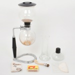 Cafeteira tipo sifão composta por dois reservatórios de vidro (superior e inferior) interligados por tubo central, manufatura da Globinho, Brasil; estrutura metálica de suporte com haste lateral e empunhadura em material sintético, tampa superior e sistema de aquecimento por lamparina a álcool com pavio. Acompanha tubo de vidro adicional, tampa da lamparina, filtro têxtil e folheto de instruções. Funcionamento por diferença de pressão: aquecimento do líquido no reservatório inferior promove a ascensão para o compartimento superior, seguido de infusão e retorno por vácuo após cessar a fonte de calor. Estado de conservação: sinais de uso; manchas internas nos reservatórios; desgaste no suporte metálico; componentes completos; funcionamento não testado, sem garantias.