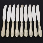 RADIO ALPACA. Conjunto de facas grandes confeccionadas em alpaca espessurado à prata, cabo de ponta oval, ricamente relevado com folhas de acanto e borda contornada por fileira perlada, lâmina em aço inox. Marca da manufatura puncionada no verso. Composto de nove(09) unidades. Apresentam marcas de uso e do tempo, vide foto. Medida: Comp.:  cm.