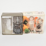 Conjunto composto por moedor de sal e moedor de pimenta, em material acrílico transparente facetado, com mecanismos internos metálicos e eixo central com botão superior de ajuste. Corpos cilíndricos com reservatório visível para grãos, bases planas e tampas superiores rosqueadas. Acompanha embalagem original litografada.