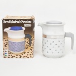 Jarra cafeteira em porcelana esmaltada, manufatura Pozzani, corpo cilíndrico com decoração geométrica aplicada por decalque, bocal vertedor lateral, alça lateral em material sintético, tampa removível e cinta metálica de fixação/ornamentação no bojo. Verso com marca de fábrica Pozzani  Made in Brazil; acompanha embalagem original litografada.