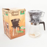 Cafeteira manual por infusão, marca Melitta, conjunto composto por jarra em vidro incolor com corpo cônico e bordo circular, munida de alça em material sintético e aro metálico de fixação; suporte filtrante em material plástico para uso com filtro de papel tipo 103. Acompanha caixa original em papelão com inscrições comerciais e material interno de acondicionamento. Inscrições: Cafeteira Manual e marca Melitta na embalagem; indicação de filtros 103.