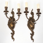 Par de arandelas de parede em bronze fundido, estilo neorrococó; espaldar recortado com composição em volutas, cartelas e motivos vegetalistas em relevo, dois braços curvos terminando em suportes para lâmpadas tipo vela, bocais com mangas simulando cera escorrida, elementos de fixação no verso com conjunto elétrico aparente. Necessita refazer parte elétrica. Estado conforme fotografias. Medidas: