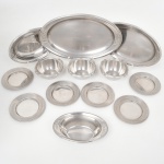 Conjunto de treze(13) peças de servir em aço inox, conformação e estampagem com relevo aplicado, composto por travessas ovais de diferentes dimensões, pratos de sobremesa com aba larga e ornamentação em relevo com motivos vegetalistas, tigelas com borda recortada em contorno ondulado e uma travessa funda, superfícies lisas com friso decorativo em relevo nas bordas;. Medidas: