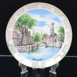 Prato em porcelana, decoração aplicada sobre esmalte,  esmaltação branca, aba larga e borda com ornamentação floral e filete em douradura, campo central com representação de vista urbana de canal em Bruges, com ponte em arco, edificações marginais e torre campanária ao fundo, inscrição BRUGGE na porção inferior, policromia aplicada; vverso com aro de apoio e cordão para suspensão. Medida: Diam.: