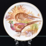 DECORAÇÃO H.N., PORCELANAS PORTUGAL. Prato de coleção em porcelana, esmaltação branca, decoração aplicada sobre esmalte, campo central com representação de faisões em paisagem campestre com vegetação, aplicada em estampagem policromada, borda circundada por friso em douradura. Verso com marca impressa em verde sob esmalte. Medida: Diam.: