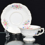 KPM - Krister Porzellan-Manufaktur, Waldenburg - Alemanha(Fundada em 1831 por Karl Kristen). Excepcional chávena para chá em porcelana de manufatura alemã, fabricada na polônia entre anos de 1930-1945, modelo Friederike, citado na Enciclopédia de Porcelana da Silésia Volume I. Série "Royal Ivory". Executado ao estilo renascentista. Esmaltagem vítrea branca, modelada com elementos fitomorfos em relevo, decorada com flores soltas em estampagem policromada, e generosos arremates de frisos em douraudura, chávena de bojo campaniforme, assente sobre base alta de formato circular e alça lateral em voluta, padrão decorativo replicado no pires. Marca da manufatura no verso. Meds.: