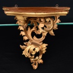 Mísula ou peanha de parede executado em madeira entalhada, com rica ornamentação de volutas, folhas de acanto e elementos vegetalistas, inspirados na estética do barroco europeu. A peça apresenta prateleira superior de contorno recortado, originalmente destinada ao apoio de pequena escultura religiosa, imagem devocional ou objeto ornamental.A estrutura inferior é composta por entalhe vazado em composição rocaille, formando conjunto de curvas e contracurvas que conferem leveza e movimento ao volume. A superfície recebeu acabamento dourado (douração ou pintura imitando ouro), com pátina escurecida em reentrâncias que realça o relevo do entalhe. Medidas: Alt.: x Comp.: Larg.: