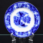 W.H. Grindley & Co., Stroke-on-Trent - Inglaterra(Fundada em 1880 por William Harry Grindley). Excepcional prato de coleção em faiança inglesa do período Vitoriano, último quartel do século XIX, c.1891. Esmaltagem vítrea na cor branca, dotada de profusa decoração na tonalidade azul cobalto, padrão "Flow Blue", conhecido pelo senso comum como "azul borrão"; centro com bela paisagem lacustre composta por personagens personagens, monumentos, edificações, imponente árvore ao centro, céu auviado e montanhas ao fundo, orla circundada por demais cenas do gênero em reserva, intercaladas por arranjos de flores e folhas de acanto, margem contornada por fileira perlada.  Marca da manufatura no verso. Med.: Diam.  cm.