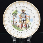 Porcelana Real, Mauá - SP(Fundada em 1943 por Fritz Erwin Schmidt). Prato de coleção em porcelana nacional da década de 1950/60, comemorativo do quarto centenário de São Paulo. Esmaltação branca, dotado de decoração central com figura de Bandeirante, aplicada em estampagem policromada, cercada pela inscrição "LEMBRANÇA DO IV CENTENARIO DE S. PAULO 1554 - 1954", borda circundada por composição geométrica em douradura. Marca da manufatura no verso. Medida: Diam.: