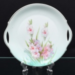 Travessa em porcelana alemã de formato circular com aba larga e duas alças vazadas laterais integradas à borda. Esmaltação branca, centro decorado com pintura floral policroma, representando hastes com flores em tons de rosa e folhas em verde, executada na técnica de decalque com realces pintados à mão. Aba com filete discreto. Verso com marca de manufatura Bavaria, indicando produção alemã. Medida: Diam.: