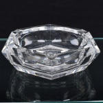 Cinzeiro em cristal incolor, de formato poligonal com bordas facetadas e espessura acentuada, apresentando quatro apoios para cigarro distribuídos na borda e cavidade central profunda. Medidas: