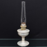 Lamparina de mesa a querosene, composta por base e reservatório em metal, queimador em latão e chaminé em vidro incolor da manufatura Alladim. Corpo com fuste central e base circular. Queimador com regulador lateral e galeria perfurada. Chaminé cilíndrica alongada, encaixada em colar metálico. Apresenta sinais de oxidação. Medidas:
