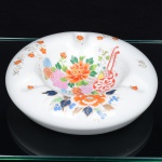 Cinzeiro em porcelana de corpo circular, com cavidade central profunda e borda larga provida de quatro entalhes equidistantes para apoio de cigarros. Decoração em esmaltação policroma sob vidrado, com composição floral estilizada em tons de laranja, verde, azul e rosa, associada a elemento ornamental de inspiração oriental. Motivos secundários distribuídos pela aba, com pequenas flores e ramos.  Fundo vazado, apresentando marca em selo vermelho com caracteres de origem chinesa. Medidas: