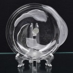 Prato decorativo em vidro incolor moldado, de formato circular com borda irregular e aba assimétrica. Corpo com relevo interno representando motivo vegetal estilizado, composto por folha nervurada e elementos orgânicos curvilíneos, com áreas parcialmente acidadas, criando contraste entre superfícies foscas e polidas. Peça apoiada em base anelar discreta, integrada ao corpo. Etiqueta original do fabricante SOGA  Japan, indicando produção japonesa. Medida: