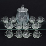 Conjunto de poncheira em cristal incolor lapidado, composto por recipiente central com tampa, acompanhado por doze(12) xícaras com alça, destinado ao serviço de ponche ou bebidas frias; corpo globular com tampa encaixada e pomo facetado, decorado com lapidação profunda composta por estrelas, raios e facetas distribuídas ao longo da superfície, xícaras apresentam forma arredondada e alça curvada, igualmente ornamentadas com motivos lapidados em padrão estrelado e raiado, criando efeitos de brilho e refração da luz. Medidas: