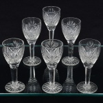 Conjunto de taças de pé alto em cristal europeu, diáfano e incolor, para licor; bojo oval dotado de profunda lapidação, contendo padrão de estrelas, sulcos, losangos e palmetas, haste sextavada, topo circundado por fileira de janelas ovais, apoiada sobre base circular, fundo preenchido por estrela raiada de dezesseis pontas. Composto de seis(06) unidades. Medidas: Alt.: 12 x Diam.: 5 cm.