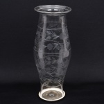 Donzela em vidro incolor, corpo alongado com leve constrição central, boca alargada com borda revirada e base circular com reservatório inferior para vela. Execução em vidro soprado em molde, com acabamento de bordo ao fogo. Decoração gravada por roda (roda de cobre) com abrasivo, composta por motivos vegetalistas com ramos, folhas e flores estilizadas, dispostos em frisos helicoidais contínuos. Medidas: Alt.: 55 x Diam.: 73 cm.