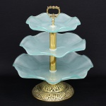 Fruteira de três andares, composta por pratos em vidro moldado de tonalidade esverdeada, com bordas onduladas, montados em haste central metálica. Estrutura em metal com banho dourado, com base circular ornada em relevo e elemento superior em forma de alça vazada com motivos vegetalistas. Um prato apresenta pequeno trincado. Medidas: Alt.: 55 x Diam.: 47 cm.