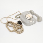 Telefone de mesa em material plástico (baquelite ou resina fenólica), de corpo monobloco com acabamento em tom cinza claro. Equipado com disco rotativo frontal numerado (sistema de discagem por pulso), com visor circular e limitador metálico lateral. Auscultador de formato anatômico, apoiado em berço superior integrado ao corpo. Cabo espiralado original, acompanhado de fiação e adaptador posterior. Modelo de fabricado entre as décadas de 1960 e 1970, amplamente utilizado em sistemas de telefonia fixa residencial e corporativa.Estado de conservação: apresenta amarelecimento do plástico, desgaste superficial, sujidades, oxidação em partes metálicas e sinais de uso. Não foi testado, sem garantias de funcionamento.