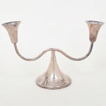 WOLFF. Castiçal de dois lumes em metal prateado, com base cônica e braços laterais curvos simétricos terminando em bocais campaniformes. Marca da manufatura puncionada ao fundo. Necessita refazer banho de prata. Medidas: Alt.: x Comp.: x Larg.: