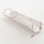 Travessa retangular em metal prateado, conformação e recorte vazado; corpo alongado curvado e laterais elevadas com borda recortada em padrão vazado com motivos repetitivos, extremidades  alças e pés integrados. Medidas: