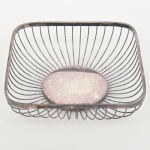 Fruteira de mesa em metal prateado, executada em estrutura de arames, com corpo de formato retangular de cantos arredondados e paredes vazadas formadas por hastes verticais arqueadas, convergindo para base central circular. Necessita polimento, apresenta desgaste no banho de prata. Medidas: