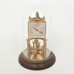 Relógio de mesa com movimento mecânico de torção, marca Schatz, com caixa em metal dourado apoiada sobre quatro colunas e base circular em madeira, protegido por cúpula em vidro. Mostrador quadrangular com numeração arábica, fundo decorado em relevo e inscrição Schatz  Made in Germany. Ponteiros metálicos centrais. Estrutura superior com elementos arquitetônicos estilizados. Mecanismo visível com pêndulo de quatro esferas giratórias (tipo anniversary), acionado por sistema de torção, montado sobre plataforma metálica circular. Não está funcionando, necessita ajustes e reparos. Medidas: Alt.: x Diam.: