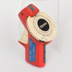 Etiquetadora manual rotativa Rotex, modelo portátil, em corpo de material plástico moldado nas cores vermelho e creme. Equipamento dotado de disco seletor circular com caracteres alfabéticos (incluindo acentuação) e numéricos, acionado por sistema mecânico de impressão por pressão, destinado à marcação de preços e identificação em etiquetas adesivas. Empunhadura anatômica integrada ao corpo, com mecanismo superior de impressão metálico. Apresenta visor/disco frontal com logomarca Rotex e escala periférica de seleção de caracteres. Laterais com plaquetas indicativas de valores, evidenciando uso comercial. Objeto típico de uso em comércio varejista, amplamente difundido na segunda metade do século XX.
