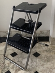 Escada dobrável de três degraus em estrutura metálica com pisos em acrílico preto. Apresenta sinais de uso, desgastes e pontos de ferrugem. Altura 63cm.