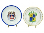 PORCELANAs, dois (2) pratos decorativos em porcelana branca, sendo: A) um (1) prato com brasão e inscrição Escola de Comando e Estado-Maior do Exército, com filete azul na borda, medindo 25cm e B) um (1) prato com borda recortada e dourada, com brasão Clube dos Embaixadores, medindo 23cm.