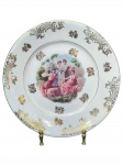 PORCELANA REAL, um (1) prato decorativo confeccionado em porcelana na tonalidade branca, decorada por cena ninfa em policromia, arranjos florais e elementos vegetalistas em vasta douração, medindo 24cm de diâmetro.