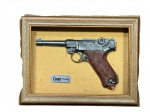 MILITARIA, uma (1) reprodução de pistola Luger P-08 confeccionada em resina, emoldurada, com plaqueta identificadora, montada sobre fundo bege e moldura de madeira, ausência do visor em vidro, medindo 24x31cm.