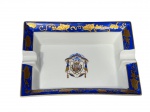 PORCELANA NG (Portugal), um (1) cinzeiro confeccionado em porcelana portuguesa na tonalidade branca, borda com faixa na tonalidade azul e guirlanda de cachos de uva e folhas de parreira em douração, centro com brasão heráldico policromado, medindo 15x11,5cm.