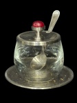 METAL PRATEADO e CRISTAL, um (1) porta geleia confeccionado em cristal translucido com tampa e presentoir em metal espessurado a prata com pega da tampa com incrustação sintétina na tonalidade vermelha, medindo 10x7cm.