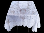 CAMA, MESA & BANHO, uma (1) toalha de mesa retangular confeccionada em linho na tonalidade branca, com bordados em tom bege, apresentando rica decoração de motivos florais, volutas, reservas centrais ovais e uma catedral gótica estilizada, como a Notre-Dame de Paris, apresenta rasgos e manchas de guardado, medindo 320x310cm.