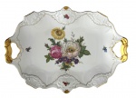 Travessa em porcelana alemã KAISER (Germany), formato oval com alças vazadas. Corpo em porcelana branca, decoração floral policromada ao centro e pequenos ramos florais distribuídos. Bordas ricamente moldadas em relevo, com acabamento em douração manual. Marca da manufatura no verso. Mede 40 x 30 cm.