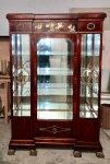 VITRINE ESTILO IMPÉRIO / NEOCLÁSSICO. Final do século XIX / início do século XX. Vitrine em madeira nobre com rico trabalho de marchetaria em padrão flameado, apresentando porta (falta a chave) central única envidraçada e laterais igualmente envidraçadas, formando três planos expositivos. Cristais frontais e laterais com bisotê. Interior com 04 prateleiras em cristal, sendo três removíveis. As janelas laterais que ladeiam a porta central recebem aplicações em bronze fundido e cinzelado, com medalhões circulares ornamentados por figuras de putti em relevo, encimados por volutas e elementos vegetalistas. No entablamento superior, friso central em bronze representando cena clássica com biga, guerreiros e cães, composição inspirada no repertório greco-romano, típica do revival neoclássico. Montantes e bases robustas, assentando sobre pés em bronze em forma de garra estilizada. Interior espelhado, ampliando a percepção de profundidade e luminosidade. Estrutura firme, sem sinais de passagem de praga. Altura: 196 cm - Largura: 123 cm - Profundidade: 43,5 cm. Atenção: ausência de chave. Encontra-se em andar térreo  Av. Atlântica / Leme  Rio de Janeiro. Retirada por conta do arrematante. Agendamento necessário pelo n. (21) 96886-7062.