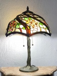 Abajur de mesa estilo Tiffany com cúpula em vitral. Elegante abajur de mesa estilo Tiffany, composto por cúpula em vitral policromado formado por múltiplos segmentos de vidro colorido unidos por molduras metálicas. Origem não identificada, mas muito possivelmente EUA. A decoração da peça apresenta motivos florais estilizados em tons de creme, verde, amarelo, vermelho e rosa, formando composição luminosa típica do repertório decorativo associado às luminárias inspiradas na produção de Louis Comfort Tiffany (Estados Unidos, final do século XIX e início do XX).A cúpula generosa em forma e tamanho é sustentada por estrutura metálica fundida em desenho orgânico, com haste central sinuosa e base circular alargada em alusão a um tronco de árvore.Sob a cúpula temos duas lâmpadas independentes com acionamento por correntes, permitindo regulagem de intensidade luminosa.Dimensões. Altura total: 55 cmDiâmetro da cúpula: 41 cmAltura da cúpula (eixo): 25 cm. Observação. Peça de inspiração Tiffany, provavelmente de produção decorativa contemporânea norte americana seguindo a técnica de segmentos de vidro colorido soldados por perfis metálicos, popularizada no movimento Art Nouveau.Estado de conservação: Sistema elétrico funcionando.Vidros e estrutura aparentam boa integridade visual nas imagens, sem trincas nem perdas. Sinais do tempo.