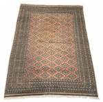 ASSINADO - Tapete oriental, possivelmente Paquistão/Bokhara, tecido à mão, lã sobre base têxtil, decoração geométrica em losangos, bordaduras ricamente trabalhadas, paleta em tons terrosos e salmão, franjas originais e assinatura visível.Medidas: 3,10 x 2,20 cm.