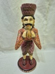 Escultura decorativa artesanal em madeira entalhada e policromada, representando figura masculina de músico Rajasthani em trajes tradicionais, com bigode característico, mãos em gesto de saudação/oferta, apoiada sobre base circular em madeira, apresentando pátina do tempo e desgastes naturais da policromia, peça de caráter étnico e decorativo. Medindo 33cm de altura. Com perda no esmalte.