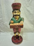 Escultura decorativa artesanal em madeira entalhada e policromada, representando figura masculina de músico Rajasthani em trajes tradicionais, com bigode característico, mãos em gesto de saudação/oferta, apoiada sobre base circular em madeira, apresentando pátina do tempo e desgastes naturais da policromia, peça de caráter étnico e decorativo. Medindo 33cm de altura. Com perda no esmalte.