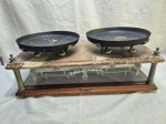 Balança antiga de duas conchas, datável do final do século XIX/início do século XX, com estrutura em metal patinado, prato superior em mármore rajado, base em madeira com mecanismo interno aparente protegido por vidro, acompanhada de duas bandejas metálicas originais e suporte auxiliar, apresentando marcas do tempo e desgaste natural compatíveis com a antiguidade. Medindo 58cm x 29cm x 16,5cm de altura.