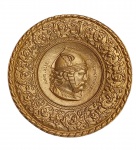 Medalhão em metal dourado Francês, Cerca de 1940. Decorado em repuxados com barrado em desenhos de arabescos no centro com perfil de efígie do chefe gaulês do povo dos arvernos e líder da revolução gaulesa contra os romanos, "Vercingetórix", ladeado por inscrições com o nome e ano "XLIX". Medidas: Diâm. 38 cm.