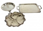 Metal espessurado a prata - Três bandejas, sendo uma retangular com alças laterais, uma circular sobre pés e uma em formato floral com relevo. Medindo: 26,5 x 16 cm; 14,5 x 2,5 cm; 15 x 16 x 3 cm