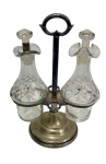 Antigo galheteiro em metal, com suporte central e alça superior, acompanhado de duas garrafas em vidro lapidado com tampas; medidas: suporte 17 x 11 x 24 cm e garrafas 7,5 x 18 cm. Necessita banho de prata.