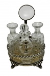 Galheteiro em metal com elegante suporte vazado, base elevada sobre pés trabalhados e haste central com alça superior, acompanhado de três garrafinhas em cristal lapidado com rica decoração geométrica, sendo duas sem tampa; medidas: suporte 17 x 11 x 24 cm e garrafas 7,5 x 18 cm. Necessita banho.