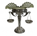 Imponente fruteira de centro composta por base em metal espessurado à prata, ricamente ornamentada com relevos florais e volutas, sustentando elegante cúpula em vidro ondulado em tom esverdeado. A peça é acompanhada por duas pequenas cestinhas laterais suspensas; Medidas da fruteira: 47,5 x 40 cm. Medidas das cestinhas: 10,5 x 15 cm.