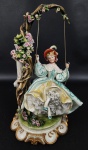 CAPODIMONTE NAPLES, Italy - Belíssima escultura em porcelana italiana esmaltada e policromada com ricos detalhes representando jovem em balanço. Peça assinada, manufatura no verso. Base com filetes em ouro. Medindo: 33 x 17 cm.
